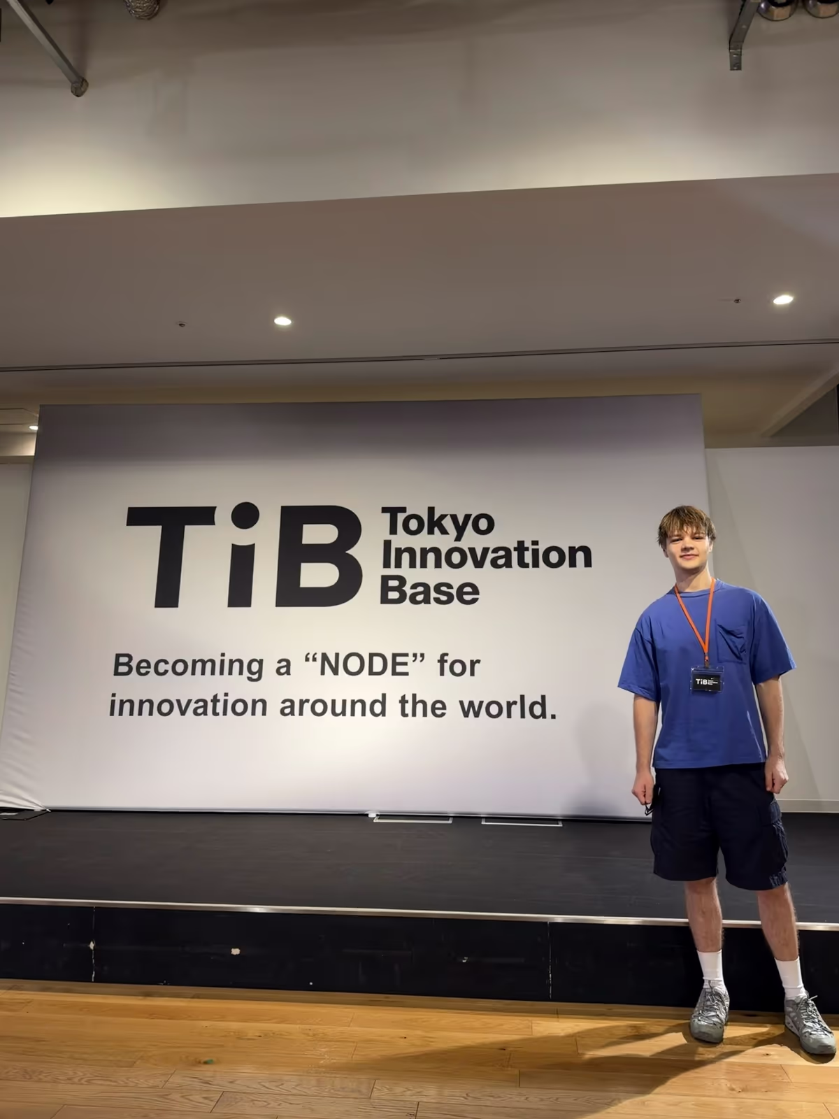 Jokubas Martysius, founder of Optify, standing in front of the Tokyo Innovation Base stage backdrop that reads 'Becoming a NODE for innovation around the world', wearing a TIB pass.